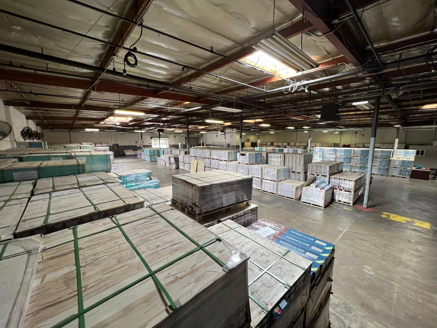 Large warehouse filled with stacked pallets of flooring materials, organised in rows under a high ceiling with exposed beams and industrial lighting.