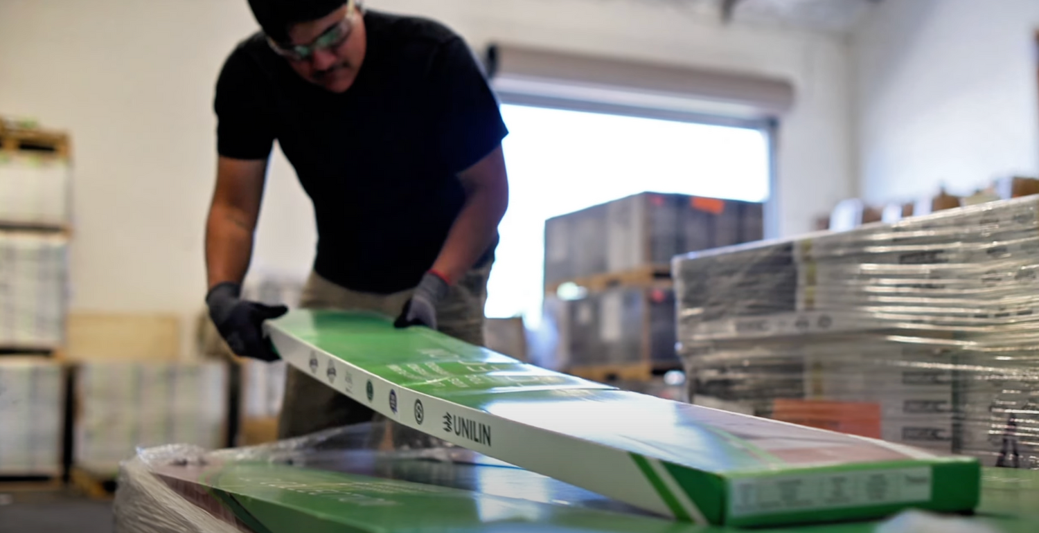 Worker handling a box of UNILIN flooring in a warehouse with stacked pallets and packaged materials in the background.
